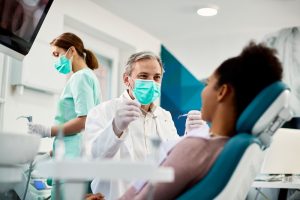 Smiling dentist with face mask talking to Black woman during dental procedure at dental clinic